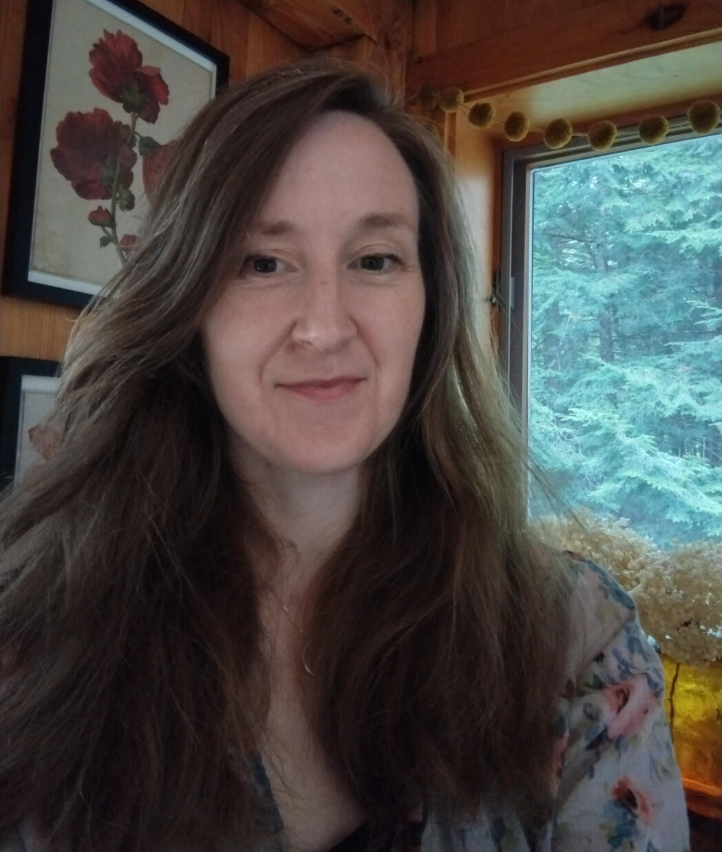A photo of Isobel Davies with a background of floral artwork and a window with forest outside.